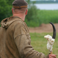 A person dressed in a brown medieval-style garment, holding a staff adorned with a carved animal skull and horn. The individual's back is turned, overseeing a lush green landscape with a lake in the background, partially obscured by trees. The scene appears to be from the 2011 Kilkim Žaibu festival.