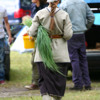 A person with long braided hair, wearing a beige shawl, grey dress, and dark apron, is seen from behind holding a large bunch of green herbs. They walk on grass towards a car with its trunk open, surrounded by others who are in casual attire and appear to be engaging in an outdoor activity reminiscent of Kilkim Žaibu 2011.