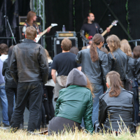 A crowd of people wearing predominantly black clothing and leather jackets attend the Kilkim Žaibu festival in 2011. Two musicians are performing on stage in the background, playing guitars. The audience stands and some sit on the grass, facing the stage, with long hair being a common feature.