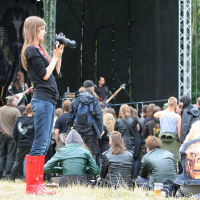 At the 2011 Kilkim Žaibu festival, a young woman with long brown hair, dressed in a black outfit and red rain boots, holds a camera and faces the stage. The crowd, some seated and others standing, watches the band perform amid trees and stage equipment. One person wears a vest with a skeletal face design.