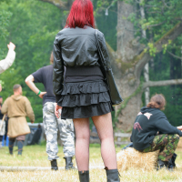 A person with vibrant red hair stands outdoors on grass at Kilkim Žaibu 2011. They are dressed in a black leather jacket, black tiered skirt, fishnet stockings, and black boots. Others are seated or standing nearby in various outfits, including camouflage pants. Trees and greenery form the background.