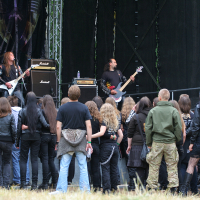 A crowd of people dressed mostly in dark clothing stands in front of a stage outdoors at Kilkim Žaibu 2011. On the stage, a band is performing with a guitarist and a bassist in the forefront. The stage has amplifiers and drum kits, and the backdrop is black with some indistinct design.