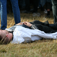 A person lies on their back on straw-covered ground at the 2011 Kilkim Žaibu outdoor event. They are wearing a white jacket over a black outfit and clasping their hands on their chest. Other people stand nearby, partially visible from the knees down. The scene appears to be in a casual environment.