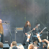 At the 2011 Kilkim Žaibu festival, a heavy metal band performs on an outdoor stage with fog effects. Three members play electric guitars and bass, wearing dark clothing, while an audience watches and raises their hands in appreciation. Amplifiers with the "Marshall" logo can be seen in the background.