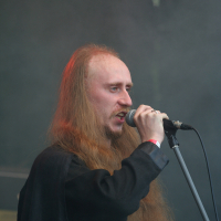 A person with long, flowing hair and a receding hairline is singing into a microphone on a stand. They are wearing a dark shirt and standing against a smoky, darkened background, likely on a stage at the 2011 Kilkim Žaibu festival. The lighting casts a slight reddish hue on their hair and face.