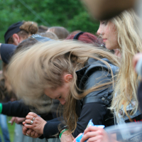 Several people stand in a row behind a metal barricade at an outdoor event. The person in the foreground has long hair flipping forward, likely headbanging. They are holding onto the barricade. Trees and greenery can be seen in the background, suggesting Kilkim Žaibu 2011 is taking place in a park.