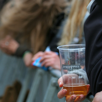 A close-up shot of a crowd at the 2011 Kilkim Žaibu festival. The focus is on a person holding a plastic cup with some beverage, possibly beer, inside it. Nearby, another person has long, blond hair flowing in the wind. Both are leaning against a metal barricade.