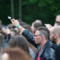A group of people stands outdoors, many holding white flowers. A man in the foreground with glasses and a leather jacket raises a white flower. The background features greenery, suggesting a wooded area. The solemn mood suggests it was a commemorative gathering, possibly during the 2011 Kilkim Žaibu event.