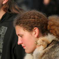 A person with curly brown hair is shown in profile, wearing a fur garment and a gold earring shaped like a star. They are looking downward, reminiscent of attendees at Kilkim Žaibu 2011. The background is blurred and out of focus, featuring other people whose details are not clear.