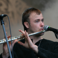 At the 2011 Kilkim Žaibu festival, a person plays a silver flute on stage into a microphone. They are wearing a black shirt and have short, dark hair. The background is foggy, creating an ethereal atmosphere. The person appears focused on their performance, with their fingers positioned on the flute's keys.