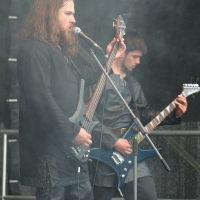 Two musicians perform on stage with smoke effects at the 2011 Kilkim Žaibu festival. The foreground musician, with long hair and a beard, sings into a microphone while playing a bass guitar. The background musician focuses on an electric guitar. Both wear black clothing, framed by a metal grid backdrop.