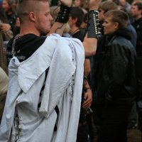 A person with short hair, dressed in a white robe with laced details and black sleeves adorned with studs, raises their hands in a gesture. They are at the 2011 Kilkim Žaibu festival, surrounded by an outdoor crowd of individuals dressed in dark clothing. Trees are visible in the background.