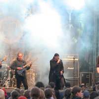 At Kilkim Žaibu 2011, a heavy metal band performs on stage amidst thick smoke. Four musicians with guitars and one vocalist stand at the front, all wearing dark clothing. Behind them, a stack of Marshall amplifiers and a drum set are visible. The crowd watches, illuminated by the stage light.