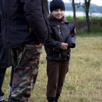 A child stands outdoors on a grassy field wearing a black beanie, brown jacket, and camouflage pants while smiling at the camera. An adult is partially visible next to the child, also dressed in outdoor gear. Trees and open landscape can be seen in the blurred background, reminiscent of a 2011 festival vibe.