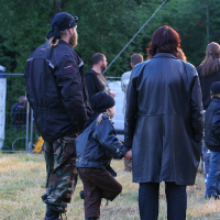 At the 2011 Kilkim Žaibu festival, a man, woman, and child, all in black jackets, stand together at an outdoor event. The man sports a bandana and camouflage pants; the woman has short hair and rolled-up jeans. The child, also wearing a bandana, holds the woman's hand while stepping on one foot. Trees and other attendees create a lively backdrop.