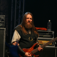 A man with long hair and a beard stands on stage, smiling while holding a brown electric bass guitar at Kilkim Žaibu 2011. He is dressed in dark clothing and wears a large ring necklace. Behind him, audio equipment and a water bottle rest atop an amplifier, with stage lighting and a partially visible crowd in the foreground.