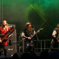 In 2011, a heavy metal band is performing on stage at Kilkim Žaibu with four male members energetically playing guitars. They are dressed in medieval or Viking-inspired attire, with long hair flowing. The stage is lit with green lighting, and fog adds to the dramatic, intense atmosphere.