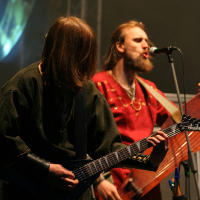 Two musicians perform on stage at the 2011 Kilkim Žaibu festival. The left musician, facing down, plays an electric guitar, with long hair partially obscuring their face and wearing a dark medieval-style outfit. The right musician, singing into a microphone, plays a string instrument and wears a red medieval-style tunic.
