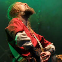 A musician with long hair and a beard passionately plays an electric guitar on stage at the 2011 Kilkim Žaibu festival. He is dressed in a medieval-style red tunic with white sleeves and ornate patterns on the cuffs. The background is lit with green stage lights, adding a dramatic effect to the performance.