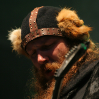 A man with a long, reddish-brown beard and mustache is playing an electric guitar. Wearing a black hat with fuzzy ear flaps and leather accents adorned with metal studs, reminiscent of a Viking helmet, he channels the energy of Kilkim Žaibu 2011. The dark background emphasizes his focused expression.