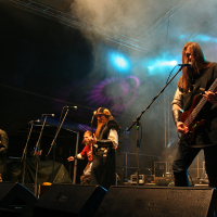 A heavy metal band performs onstage under colorful lights at Kilkim Žaibu 2011. The musicians wear medieval-inspired outfits; one plays an electric guitar, another a bass guitar, and another sings into a microphone. A drummer is visible in the background. Smoke and dark lighting set a dramatic atmosphere.