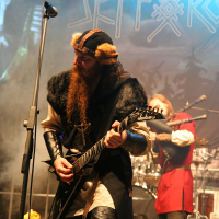 A musician with a long red beard, dressed in a Viking-inspired outfit, plays an electric guitar on stage. Behind him, another performer in a red and black costume plays drums. The stage backdrop features the name "Skálmöld" with Norse motifs. Smoke and blue lighting enhance the dramatic setting at Kilkim Žaibu 2011.