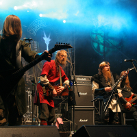 Four musicians perform on stage with guitars, dressed in medieval-themed costumes at Kilkim Žaibu 2011. Blue stage lighting and fog create a dramatic atmosphere. Amplifiers and a drum set are visible in the background. The backdrop displays band logos and artwork. The musicians are actively playing their instruments.