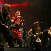 Musicians perform on a dimly lit stage at the 2011 Kilkim Žaibu festival. Four guitarists are playing: one in black, one in green, one in red (also playing a flute), and one in black with a fur collar. Amplifiers and drums stand visible in the background, accompanied by a backdrop of intricate designs.
