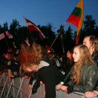 A crowd at the 2011 Kilkim Žaibu festival enthusiastically listens to live music. Attendees, many with long hair and dressed in dark clothing, lean against a metal barricade, some headbanging. Several flags, including the Lithuanian flag, are held aloft amidst the dusk setting with trees in the background.