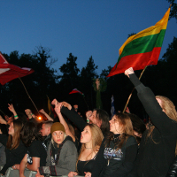 A crowd at the Kilkim Žaibu 2011 event raises hands and flags, including a tricolor flag of green, yellow, and red. It's evening with a dusky blue sky. The audience, dressed predominantly in dark attire, is enthusiastic, with people singing along and waving flags amidst trees in the background.