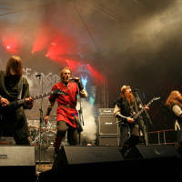 A heavy metal band performs on an outdoor stage at the 2011 Kilkim Žaibu festival. Four musicians in dark, medieval-inspired outfits rock with guitars under vibrant red and blue lights. The background features a large band logo. Amplifiers and speakers are visible behind them, and mist adds to the dramatic atmosphere.