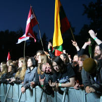 A crowd of excited concertgoers stands behind a metal barrier at the 2011 Kilkim Žaibu festival. Many are raising their hands in the air and waving flags, including a Latvian flag and a Lithuanian flag. The event appears lively, with people smiling and cheering under a dark night sky.