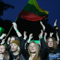 A group of concertgoers stands with their arms raised in the air at an outdoor event during nighttime at the 2011 Kilkim Žaibu festival. Some are clapping, and most appear engaged with the performance. One person is holding up a large flag with horizontal stripes of yellow, green, and red in the background.