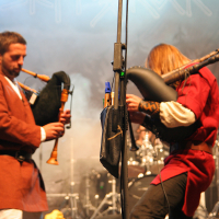 At the 2011 Kilkim Žaibu festival, two musicians performing on stage both play bagpipes. The musician on the left wears a brown tunic with white sleeves, while the musician on the right dons a red tunic. A drum set is visible in the background, along with some stage lights and smoke effects.