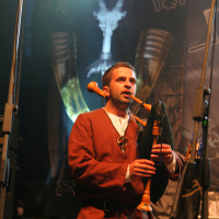 A man is playing a bagpipe on stage, dressed in medieval clothing at the 2011 Kilkim Žaibu festival. He stands between two microphones with a dark backdrop featuring a large logo, two horns, and decorative elements illuminated by bright stage lights. Musical equipment and cables are visible on the floor.