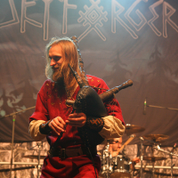A man in a red tunic plays bagpipes on stage at the Kilkim Žaibu 2011 concert. Behind him, "Skyforger" is displayed on a large banner. The stage is dimly lit with blue and yellow spotlights, and a drummer is partially visible in the background.