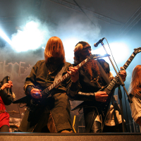 At the 2011 Kilkim Žaibu festival, a heavy metal band performs on stage with dramatic lighting and fog effects. The musicians, dressed in medieval-style outfits, passionately play their instruments, including electric guitars and bagpipes. The scene is energetic and intense, capturing the raw emotion of the performance.