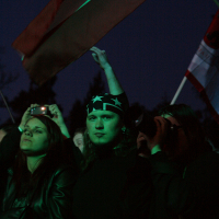 A crowd at an outdoor event, mostly in dark clothing. One person in the center wears a bandana with star patterns. Another person nearby holds a flag, partially illuminated by event lighting, creating a dramatic effect against the night sky. Expressions are varied, showing attentiveness at Kilkim Žaibu Festival 2011.