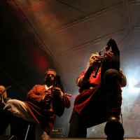 At the 2011 Kilkim Žaibu festival, two musicians perform on stage under dim lighting. The central focus is on one musician playing bagpipes and wearing a red tunic, while the other, also playing bagpipes, is dressed in a brown outfit. Smoke and colorful stage lights create a dramatic atmosphere. The audience is not visible.