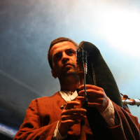 A man in a brown tunic plays the bagpipes at the 2011 Kilkim Žaibu festival, holding the chanter with both hands. A foggy, misty atmosphere surrounds him, with a bright light source illuminating from behind, creating a dramatic effect. His facial expression is focused and intense as he performs.