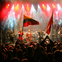 A rock band performs on stage with a large, enthusiastic crowd in the foreground at Kilkim Žaibu 2011 festival. The stage is illuminated with colored lights, and flags from various countries are being waved by audience members. The band's backdrop features their logo in bold letters.