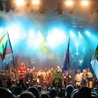 A vibrant concert stage at the 2011 Kilkim Žaibu festival features performers in medieval costumes holding various national flags. Bright lights and fog create a dramatic atmosphere. The audience, visible in the foreground, watches as the performers stand in a row, some raising their hands or flags in celebration.