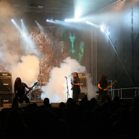 At Kilkim Žaibu 2011, a heavy metal band performs energetically on a smoke-filled stage. The guitarist on the left swings his hair, while the lead singer holds the microphone at the center. The bassist on the right plays intensely. Bright stage lights and a backdrop with band graphics create a dramatic atmosphere.