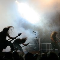 Three musicians with long hair perform on a dimly lit, smoky stage at Kilkim Žaibu 2011. They energetically play electric guitars under a bright white spotlight, with one microphone stand visible in the center. The audience is partially visible in the foreground.