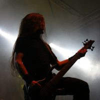 A musician with long, flowing hair plays a black electric guitar on stage at the 2011 Kilkim Žaibu festival. He is dressed in all black and surrounded by dramatic stage lighting, creating a high-contrast silhouette effect. Smoke and bright beams of light in the background add to the atmospheric, concert-like setting.