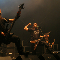 At the 2011 Kilkim Žaibu festival, a heavy metal band performs on stage, featuring three members. The bassist on the left plays with intensity, the lead vocalist in the center shouts into a microphone while raising his fist, and the guitarist on the right focuses on his instrument. The stage is dimly lit, with amps and equipment visible in the background.