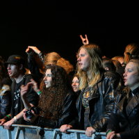 Concert-goers with long hair and leather jackets stand behind a metal barricade, enthusiastically watching a performance at the Kilkim Žaibu festival in 2011. Some have their hands raised, and one person is holding a camera. The dark background emphasizes the crowd's excited expressions and gestures.
