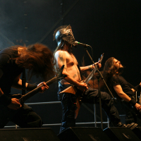 At Kilkim Žaibu 2011, four musicians perform energetically on stage. The shirtless lead singer wears a helmet and sings into a microphone. All band members have long hair and play electric guitars. The stage background is dark with stage lights illuminating the performers from above, creating a dramatic effect.