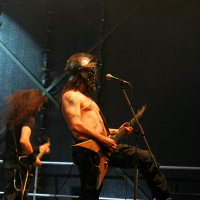 Two musicians performing on stage, with the focus on a shirtless guitarist in the foreground wearing a medieval-style helmet. The guitarist is playing an electric guitar and standing near a microphone. To the left, another musician with long, curly hair is playing a guitar against a dark background at Kilkim Žaibu 2011.