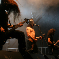 Three musicians perform on stage during the heavy metal concert at Kilkim Žaibu 2011. The guitarist on the left has long hair obscuring his face, headbanging. The shirtless vocalist in the middle wears a black mask with horns and sings into a microphone. The guitarist on the right plays intensely amid smoky effects.